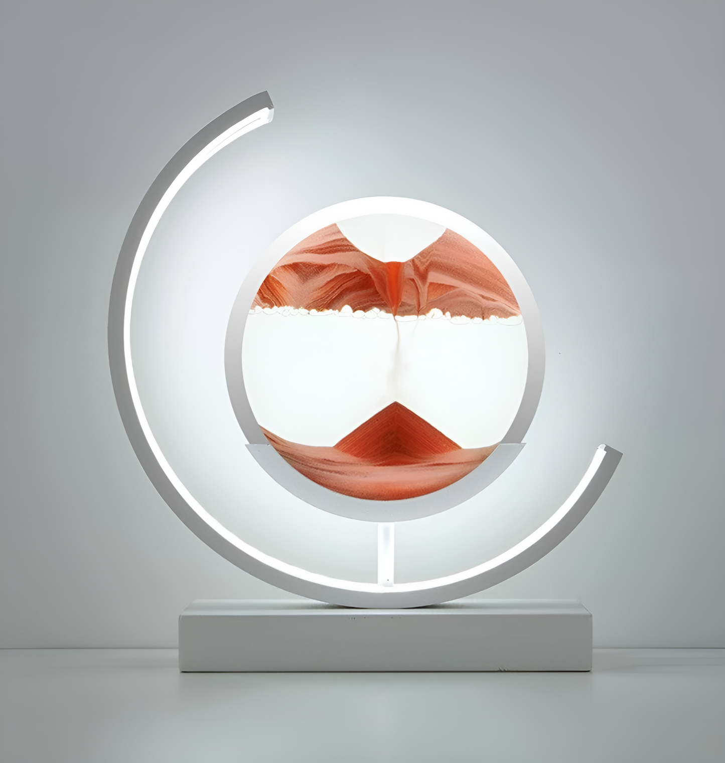 De afbeelding toont een stijlvolle LED-lamp met een uniek zandkunstontwerp in het midden. Het zand is verdeeld in zachte tinten van oranje en wit, die samen een bergachtig landschap creëren. De lamp wordt omlijst door een gebogen LED-licht dat een warme, uitnodigende gloed uitstraalt, perfect voor het creëren van een sfeervolle en moderne uitstraling. De strakke, witte basis maakt het geheel af, wat deze lamp tot een elegante toevoeging maakt voor elk interieur.
