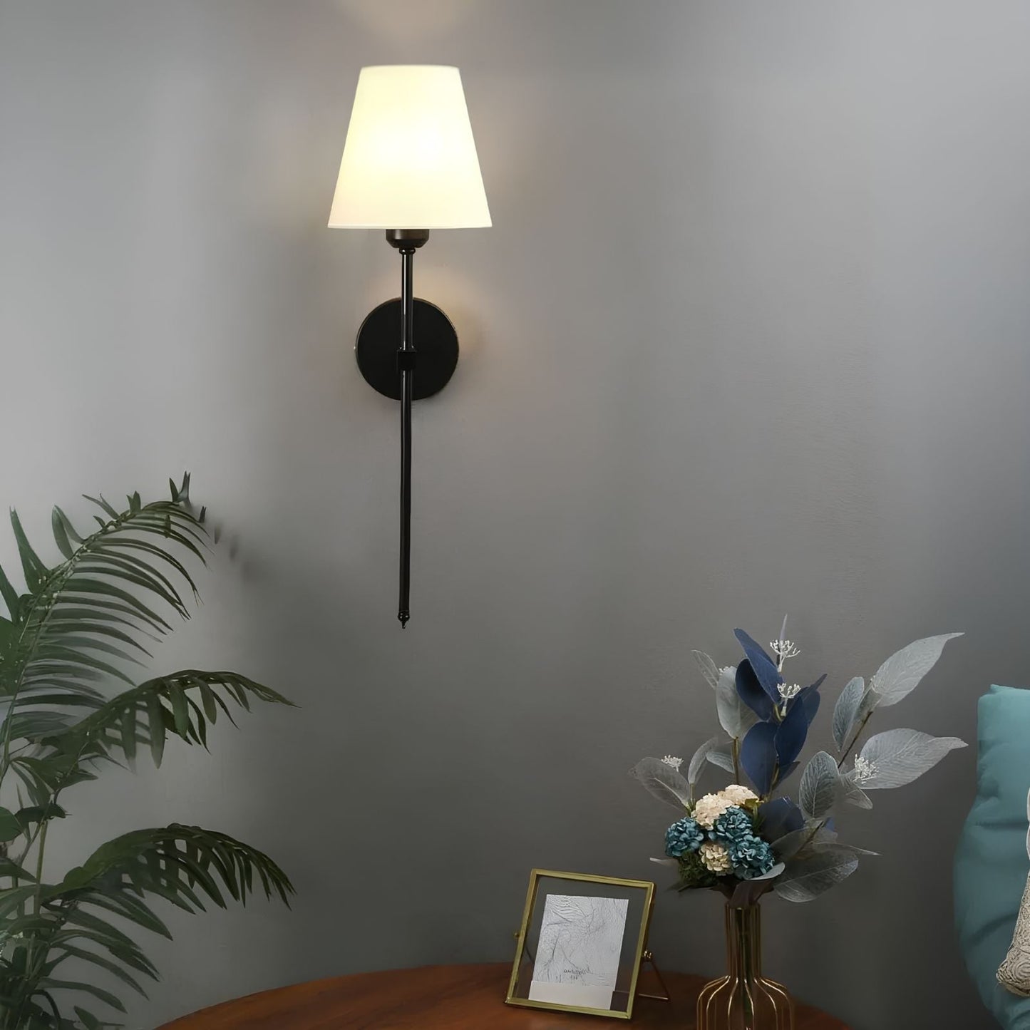 De Aurelia Wall Light met een klassieke zwarte afwerking en een witte lampenkap verlicht een stijlvolle ruimte. De wandlamp is bevestigd aan een grijze muur en verfraaid met een plant, bloemen en een ingelijste tekening op een houten tafel, waardoor een warme en verfijnde sfeer ontstaat.