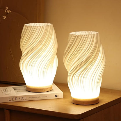 Twee Balaara Serene Wavecrest lampen met golvende patronen, stralen warm wit licht uit op een houten tafel met een boek ernaast, perfect voor een kalme en rustgevende sfeer.