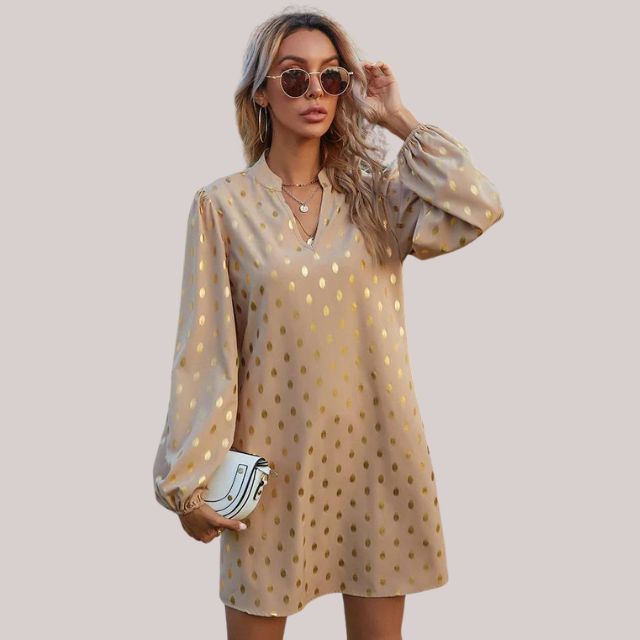 De Bellara Luxe jurk, een elegante beige jurk met glanzende gouden details en lange pofmouwen. Deze verfijnde jurk is perfect voor een chique uitstraling en ideaal voor feestelijke of formele gelegenheden