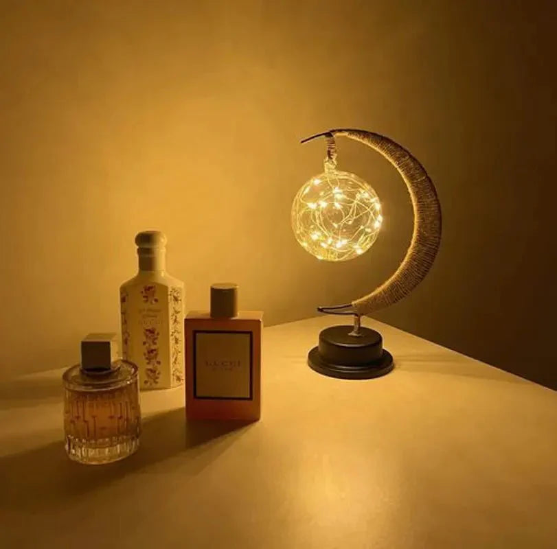 Een betoverende maanlamp die een magische gloed verspreidt, verlicht door delicate lichtjes in een gloeiende bol. De lamp staat op een elegante zwarte basis en wordt omringd door luxe parfumflessen, wat een luxe en serene sfeer creëert. Perfect voor een stijlvolle en warme ambiance in elke ruimte.