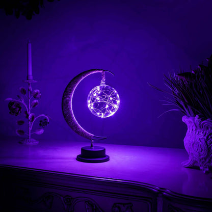 Een magische maanlamp verlicht met paarse LED-lichten, die een dromerige sfeer creëert. De lamp bevat een glinsterende bol van lichtjes, ondersteund door een elegante halve maanstructuur op een zwarte basis. De verlichting is perfect voor een mystieke, kalmerende ambiance in elke kamer.