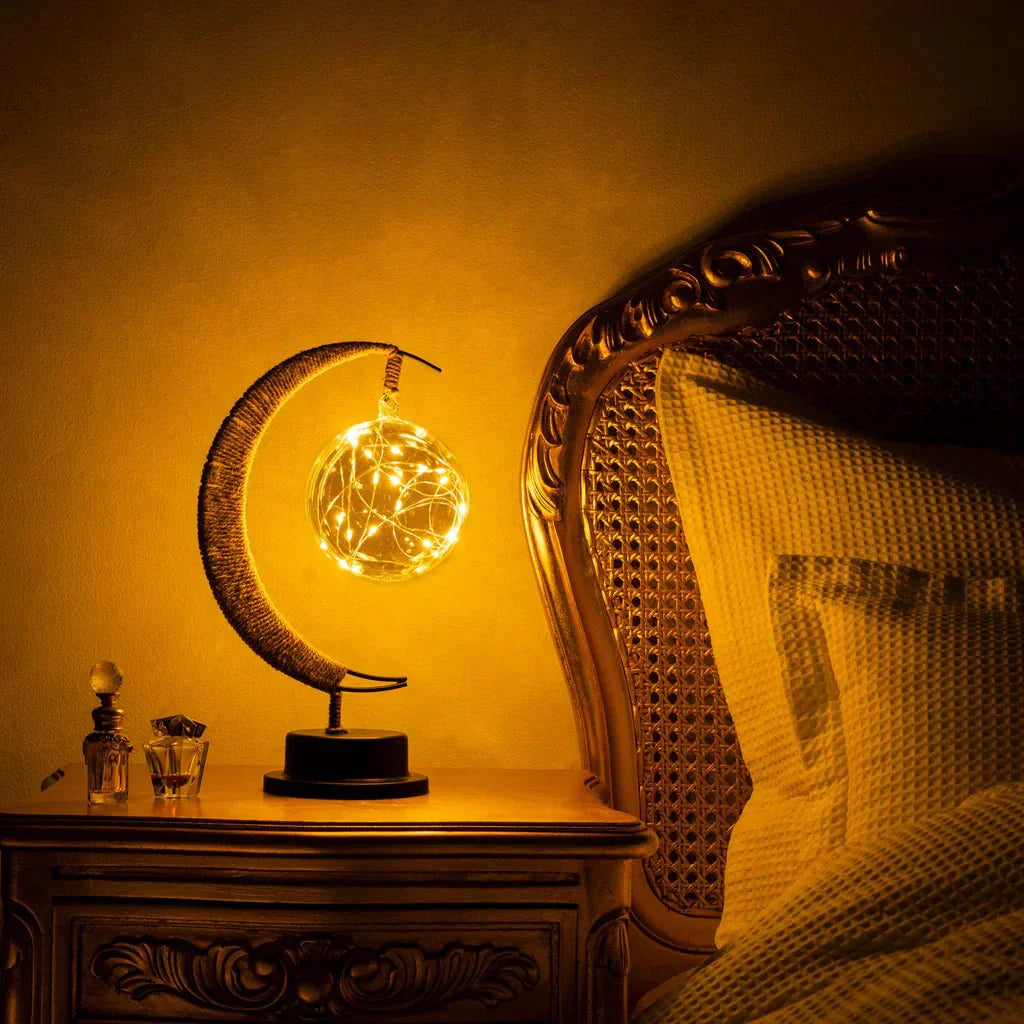 De GEMYNORA maanlamp, verlicht met warme, gloedvolle LED-lichtjes, creëert een magische sfeer op een nachtkastje naast een bed. De lamp is ontworpen met een elegante halve maan en een stralende bol van lichtjes, wat zorgt voor een dromerige en rustgevende ambiance in de kamer.
