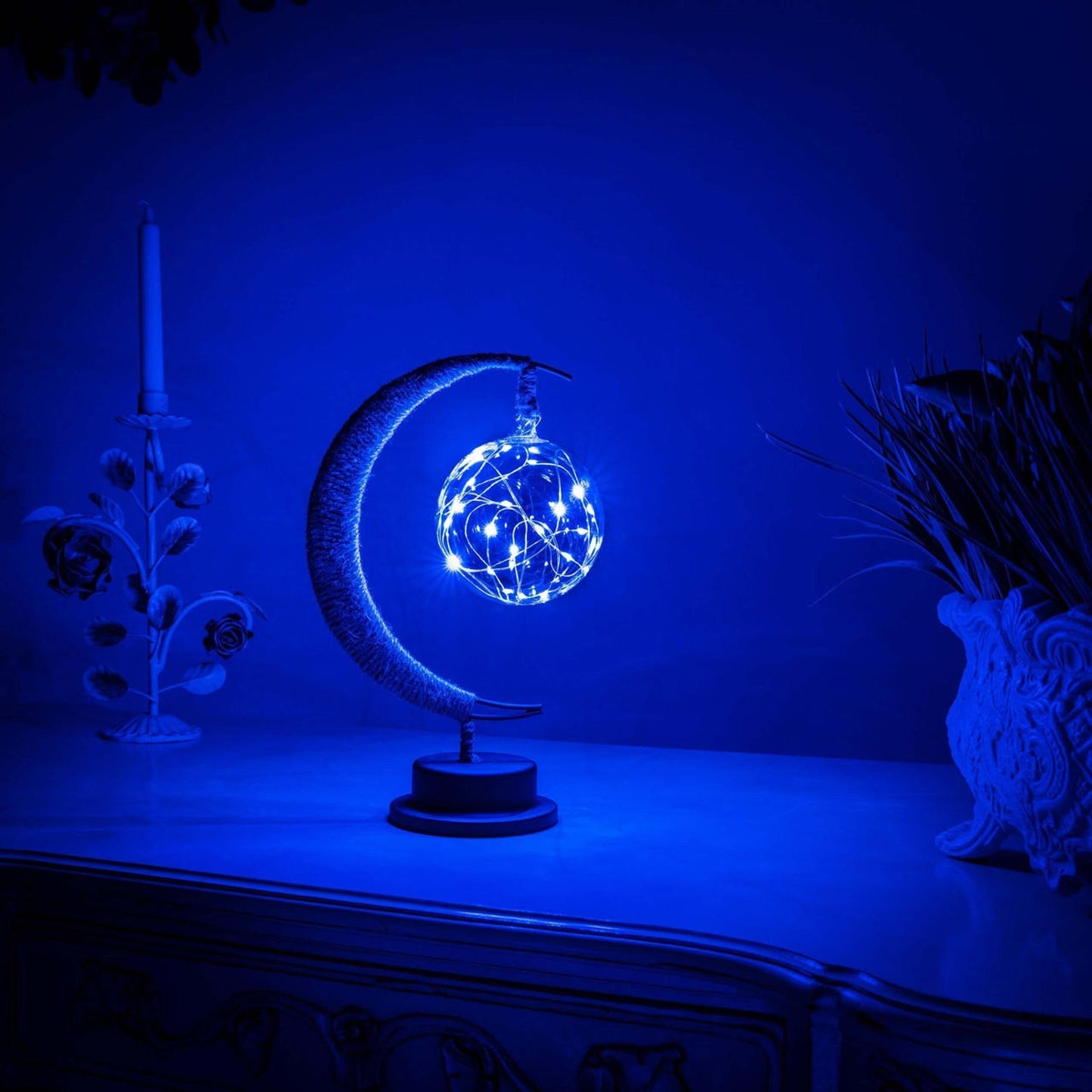 De GEMYNORA maanlamp, met een betoverende blauwe gloed, verlicht een nachtkastje in een rustige kamer. De lamp, met een halfmaansvorm en een stralende bol van lichtjes, creëert een magische sfeer en voegt een dromerige, kalmerende uitstraling toe aan het interieur.