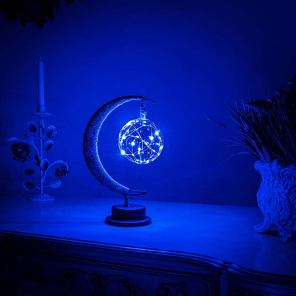 De GEMYNORA maanlamp, met een betoverende blauwe gloed, verlicht een nachtkastje in een rustige kamer. De lamp, met een halfmaansvorm en een stralende bol van lichtjes, creëert een magische sfeer en voegt een dromerige, kalmerende uitstraling toe aan het interieur.