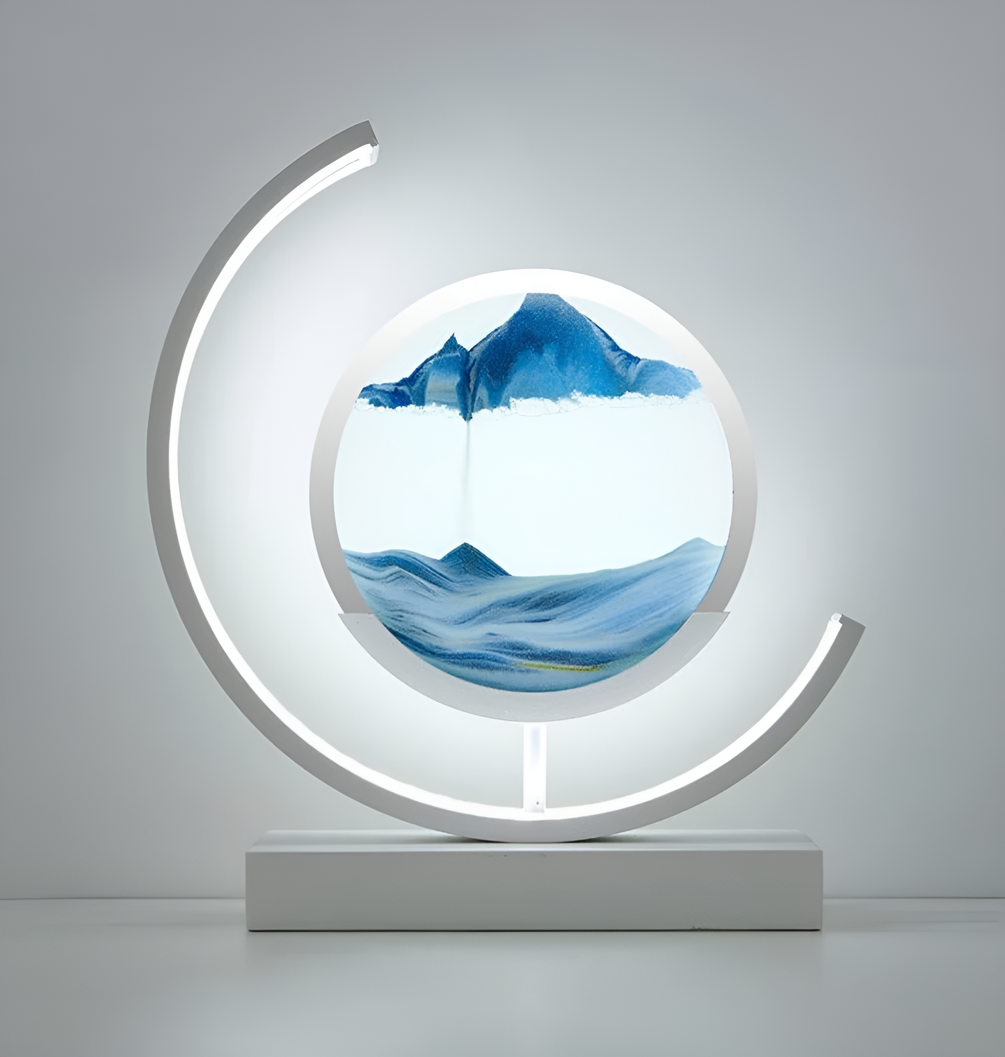 De afbeelding toont een stijlvolle LED-lamp met een dynamische ronde zandkunst in het midden. Het verlichte ontwerp bevat zachte blauwe en witte tinten in het zand, die samen een rustgevend landschap creëren. De lamp is omgeven door een gebogen LED-licht dat een warme gloed uitstraalt, waardoor het een moderne en elegante uitstraling krijgt. De witte basis van de lamp zorgt voor een strakke afwerking, ideaal voor een eigentijdse interieurstijl.