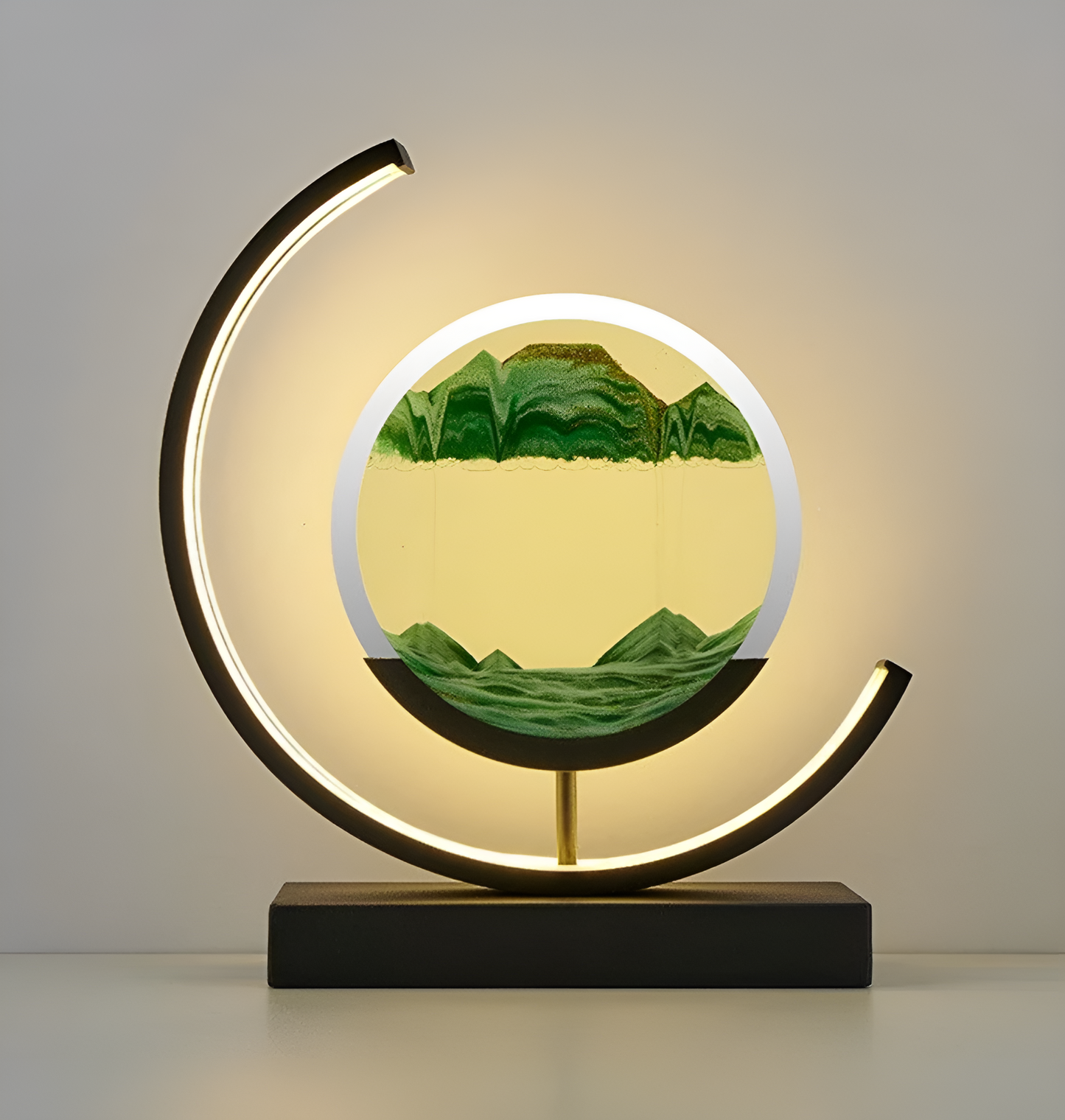 De afbeelding toont een elegante LED-lamp met een unieke ronde zandkunst in het midden. Het zand is in verschillende tinten groen en geel, wat een bergachtig landschap simuleert. De lamp wordt omlijst door een gebogen LED-licht dat een warme gloed uitstraalt, perfect voor het creëren van een sfeervolle en moderne ambiance. De zwarte basis van de lamp biedt een strakke en verfijnde afwerking, waardoor deze lamp een stijlvolle aanvulling is op elk interieur.