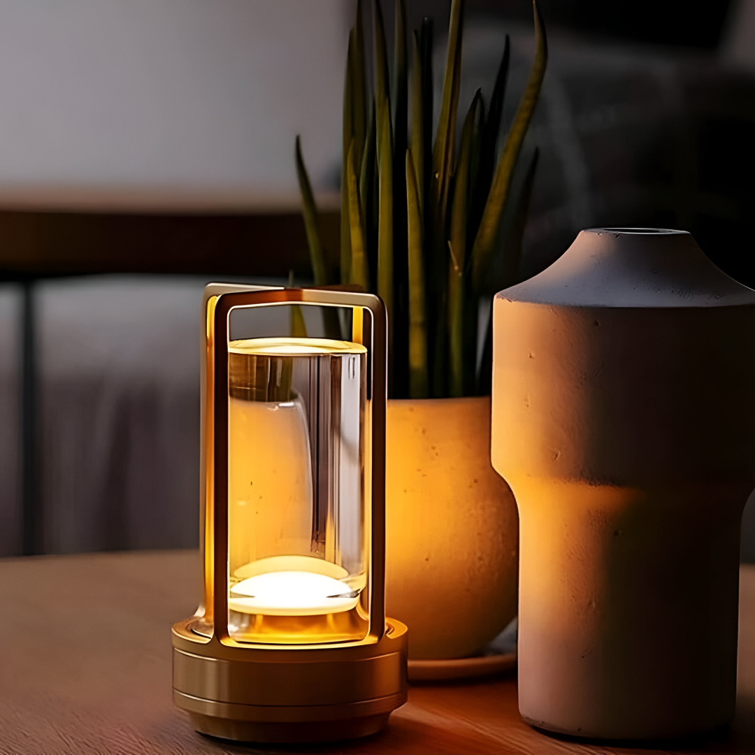 Een stijlvolle, goudkleurige, waterbestendige lantaarnlamp staat op een houten oppervlak naast een potplant en een keramieken vaas en gloeit zachtjes in een schemerige kamer. De lamp straalt een warm licht uit en creëert een gezellige sfeer.
