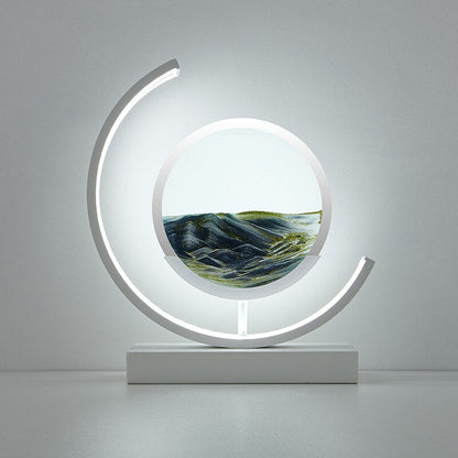 De LumiAura lamp toont een rustgevend zandlandschap in zachte groene tinten, met de zandlagen die langzaam bewegen, verlicht door het omringende zachte licht. Het strakke, moderne ontwerp voegt een kalmerende sfeer toe aan elke kamer, perfect voor ontspanning. Het kunstzinnige zandlandschap creëert een betoverende uitstraling, waardoor het een perfecte aanvulling is voor je interieur.