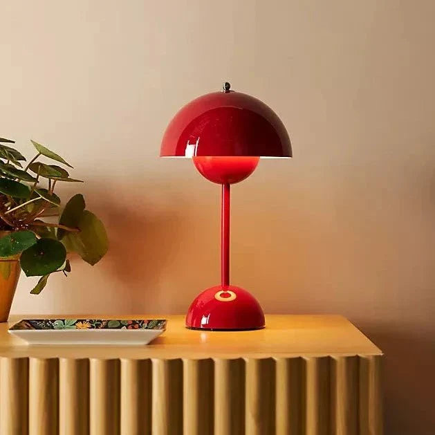 Een moderne rode LumiNova tafellamp met een elegante bolvormige kap, geplaatst op een houten tafel naast een decoratieve schaal en een groene plant. Deze stijlvolle LED-lamp biedt sfeervolle verlichting en is perfect als opvallend accessoire voor woon- of werkruimtes.