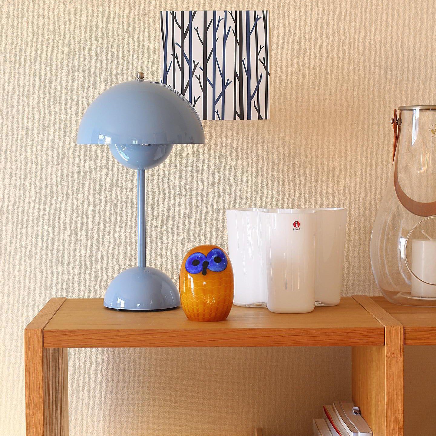 Een elegante blauwe LumiNova tafellamp met een bolvormige kap, geplaatst op een houten plank naast een decoratief glazen uiltje en een paar witte glazen. De lamp biedt stijlvolle LED-verlichting en is een perfect accent voor elke moderne en sfeervolle inrichting.