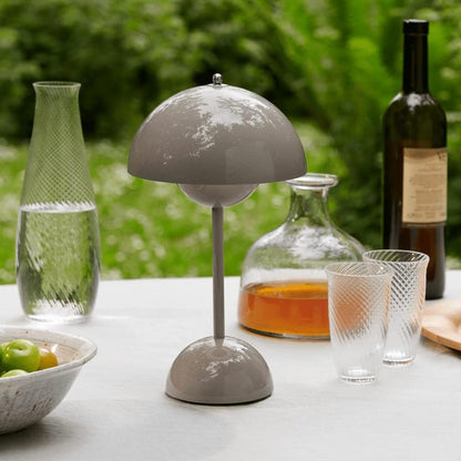 Een stijlvolle LumiNova tafellamp in grijze kleur, geplaatst op een buitentafel naast glazen drinkbekers en een karaf met drank. De lamp biedt elegante LED-verlichting en voegt een verfijnd accent toe aan de buitenruimte, ideaal voor een gezellige sfeer.