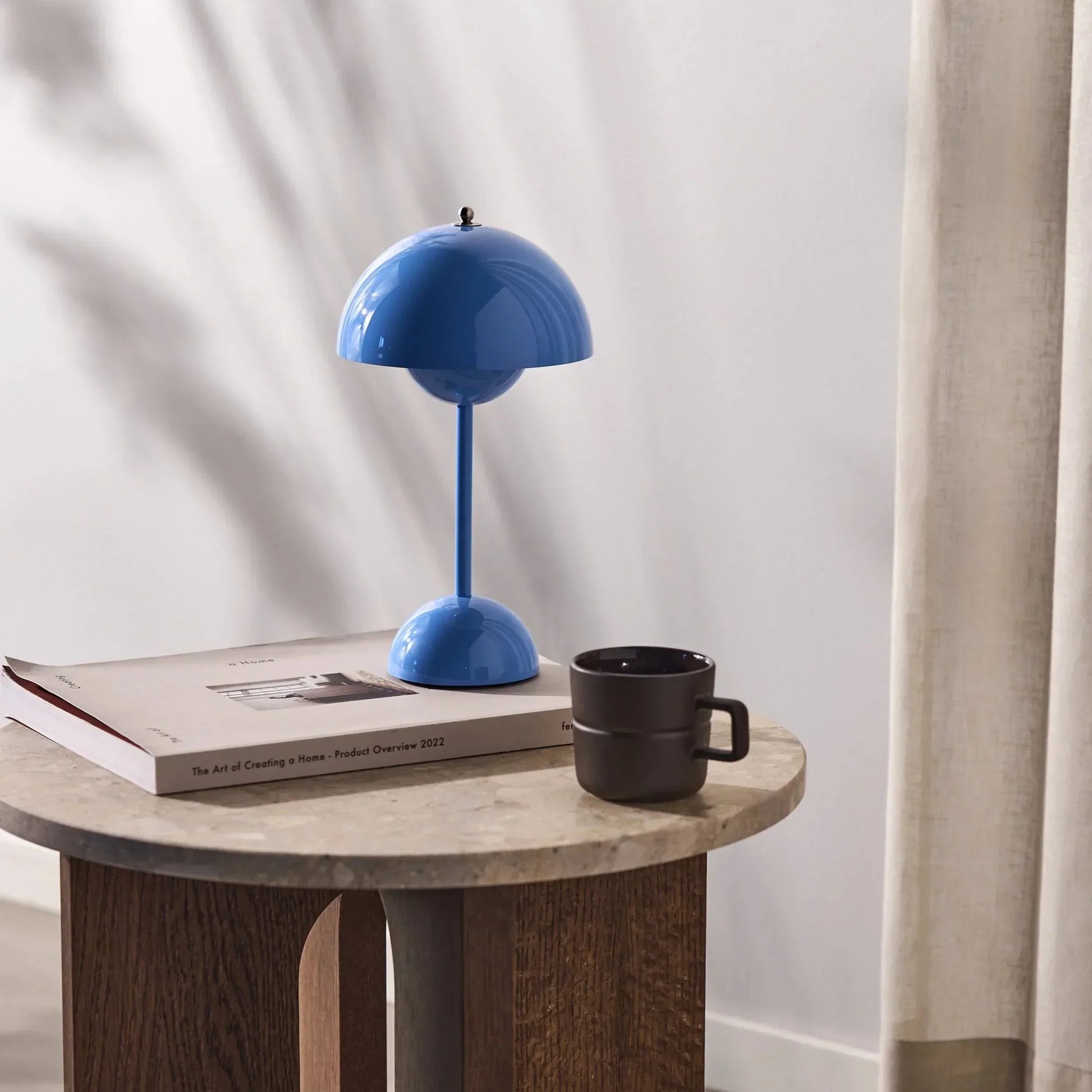 Een heldere blauwe LumiNova tafellamp op een houten salontafel, naast een koffiemok en een stijlvolle boek. Deze moderne LED-lamp biedt stijlvolle verlichting en is een opvallend en verfijnd accessoire voor elke woonkamer of werkruimte.