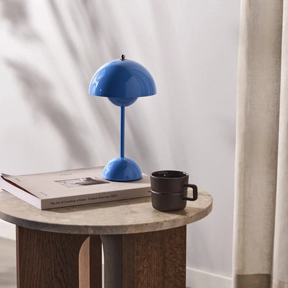 Een heldere blauwe LumiNova tafellamp op een houten salontafel, naast een koffiemok en een stijlvolle boek. Deze moderne LED-lamp biedt stijlvolle verlichting en is een opvallend en verfijnd accessoire voor elke woonkamer of werkruimte.