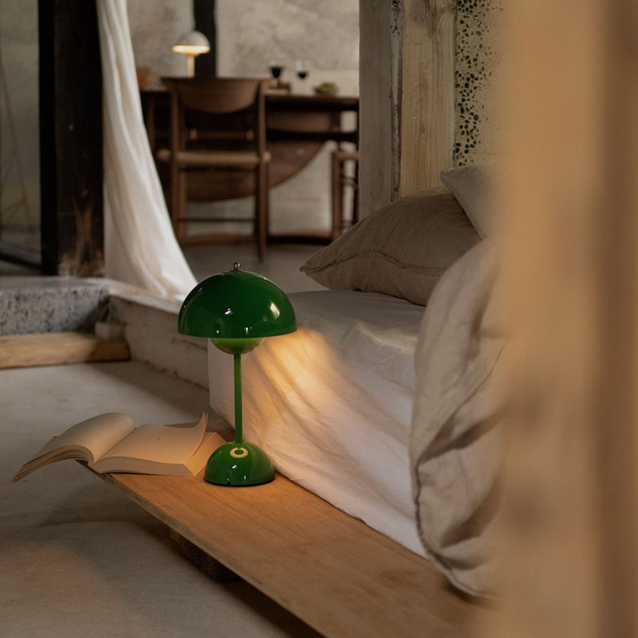 Een sfeervolle slaapkamer met een LumiNova tafellamp LED in groene kleur, geplaatst naast een bed. De lamp verlicht zachtjes een open boek dat op de houten vloer ligt, waardoor een gezellige en rustgevende sfeer ontstaat in de kamer. De stijlvolle en moderne tafellamp biedt de perfecte verlichting voor ontspanning en lezen.
