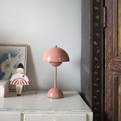 Een charmante kamer met een LumiNova tafellamp LED in een zachte roze kleur, geplaatst op een witte kast. De lamp straalt een warme, uitnodigende gloed uit en vormt een elegant contrast met de kunst en het houten kastmeubel in de achtergrond. De minimalistische en moderne lamp is een perfecte aanvulling op het interieur, die zowel stijlvol als functioneel is.