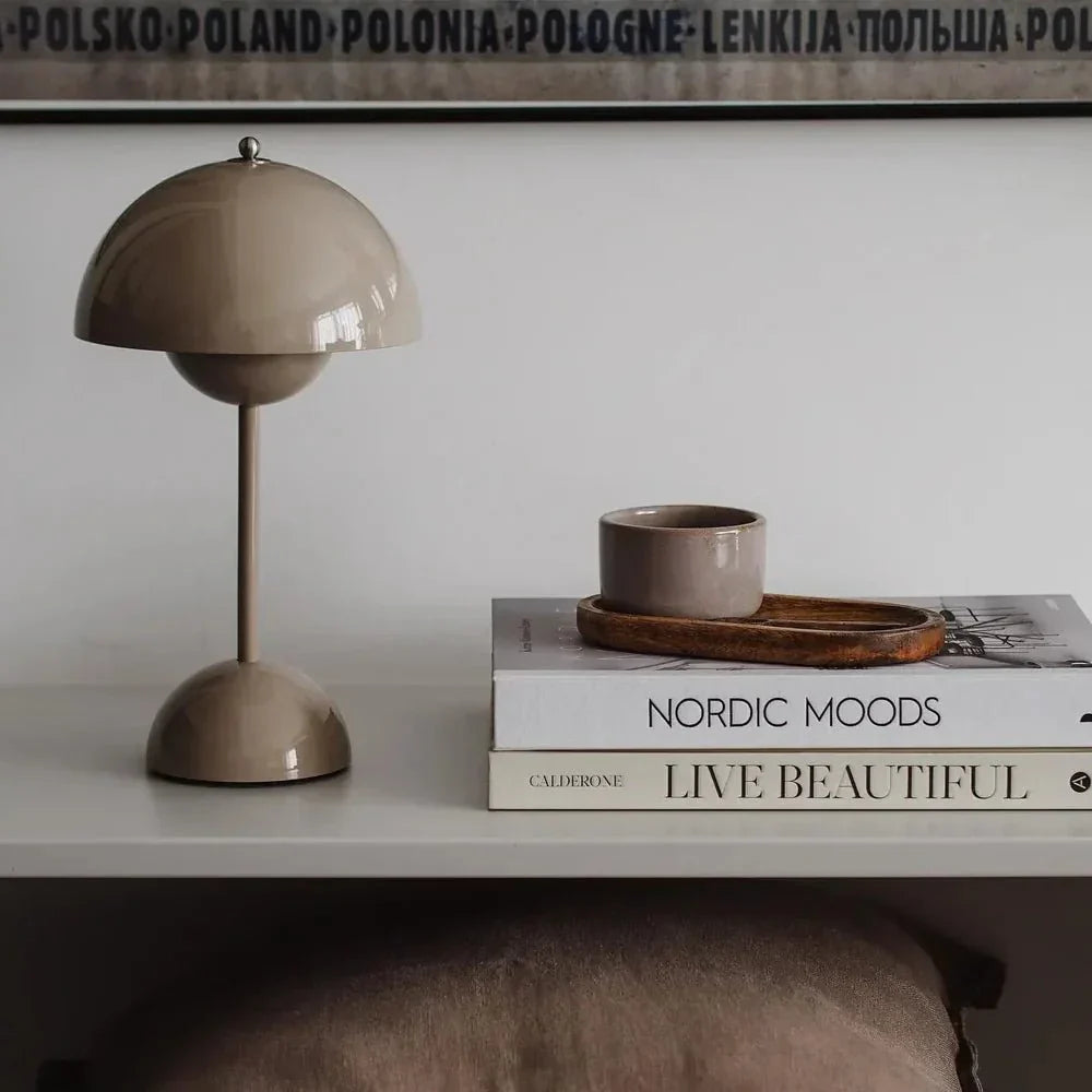 Een elegante LumiNova tafellamp in een zachte beige tint, geplaatst naast een klein keramisch kopje op een stapel boeken. De lamp heeft een iconische halfronde kap en een slanke, minimalistische voet, perfect voor het toevoegen van stijlvolle verlichting aan elk modern interieur.