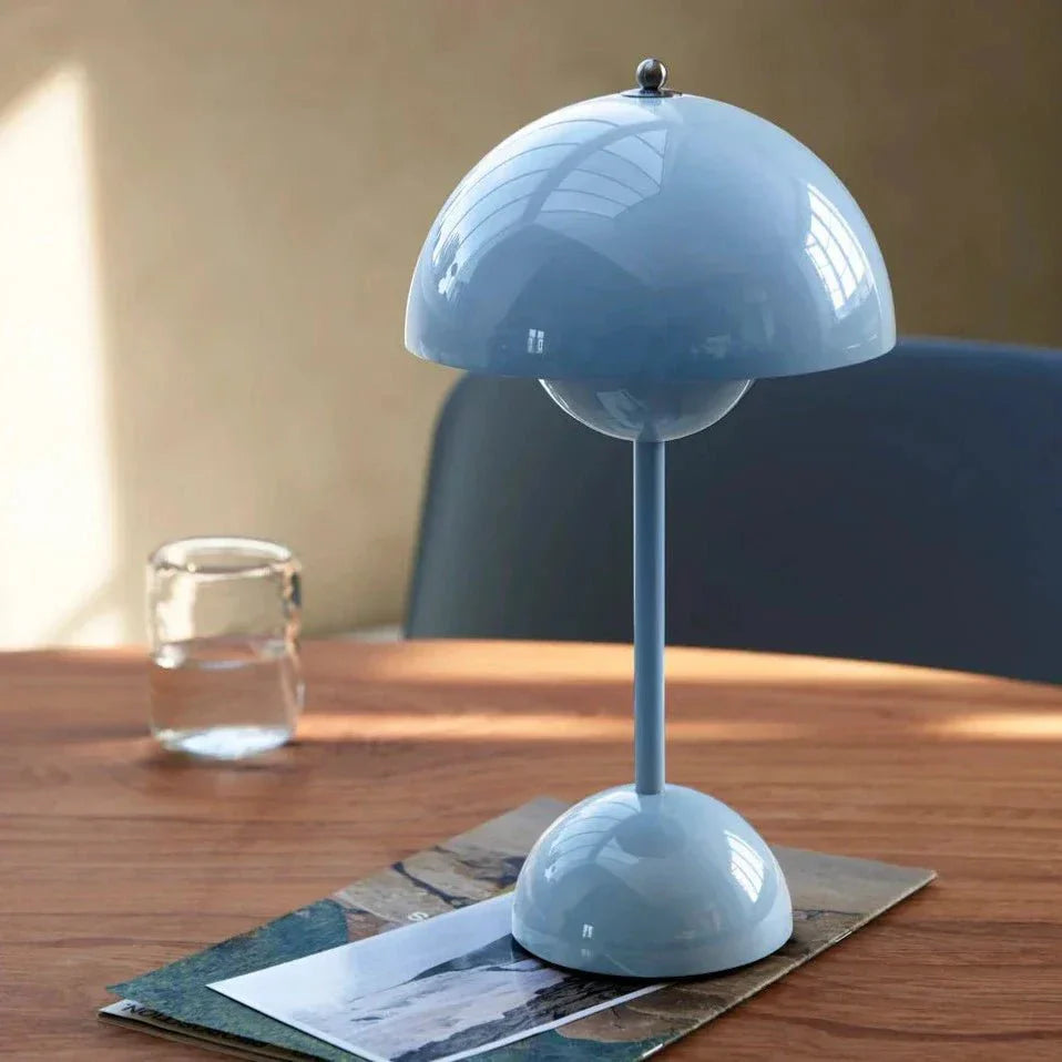 Stijlvolle LumiNova LED-tafellamp in lichtblauw, geplaatst op een houten tafel met een glas water en tijdschriften, ideaal voor sfeervolle binnenverlichting.