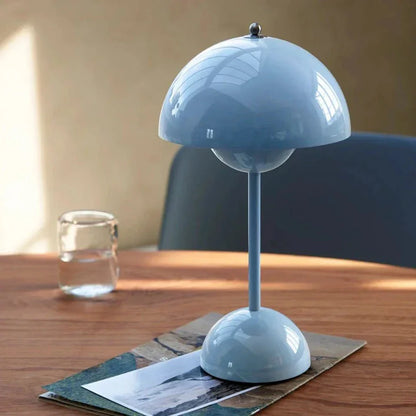 Stijlvolle LumiNova LED-tafellamp in lichtblauw, geplaatst op een houten tafel met een glas water en tijdschriften, ideaal voor sfeervolle binnenverlichting.