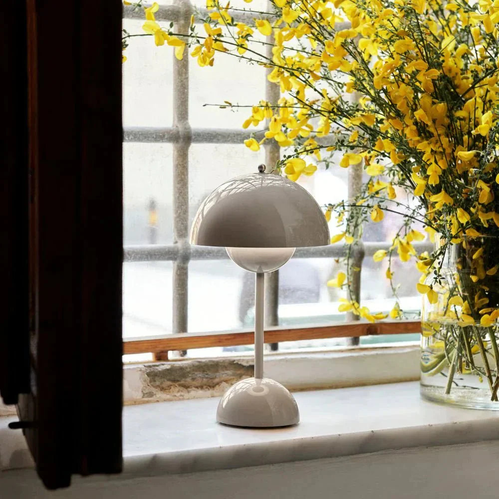 De LumiNova LED-tafellamp in een elegante beige tint, gepresenteerd op een vensterbank naast een vaas met levendige gele bloemen, met uitzicht op een rustieke achtergrond.