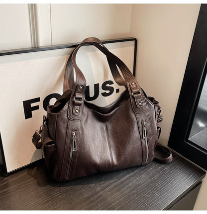 OLYNDA tote bag in donkerbruin kunstleer, met twee ritsvakken aan de voorkant, stevige handgrepen en een schouderriem. Stijlvol en praktisch ontwerp, geplaatst naast een 'FOCUS' bord.