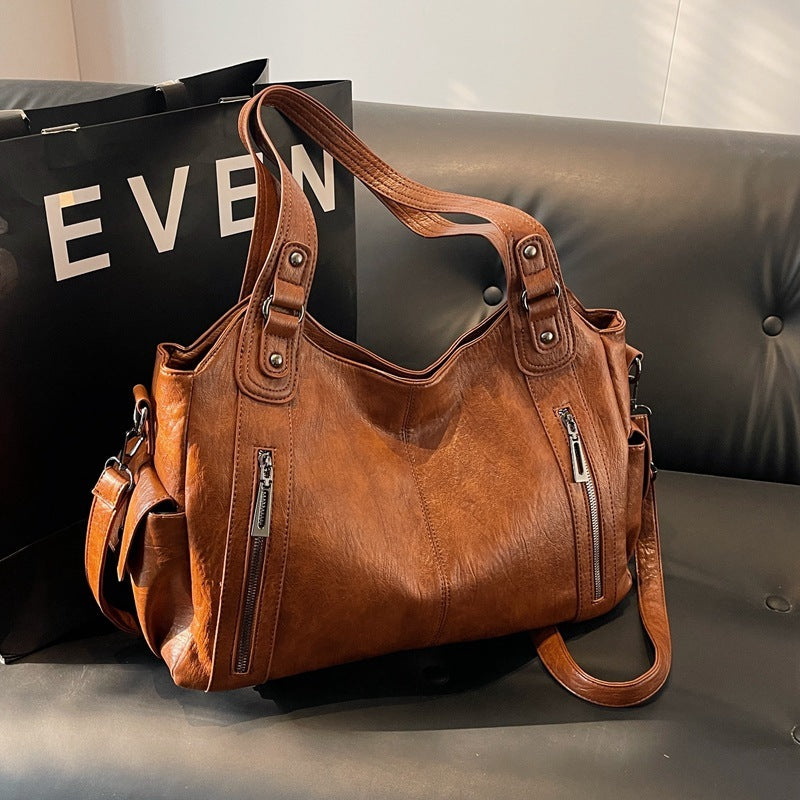OLYNDA bruine leren tote tas met meerdere ritsen, ideaal voor dagelijks gebruik, geplaatst op een zwarte leren bank met een stijlvolle uitstraling.