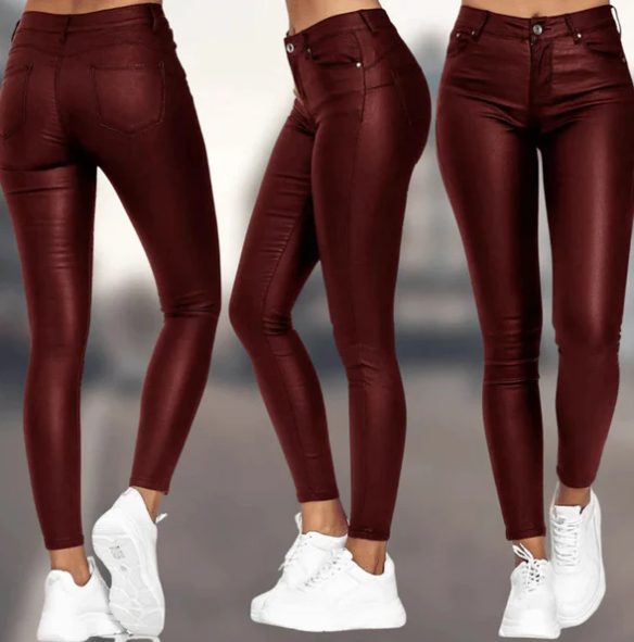 Een vrouw draagt een bordeauxrode, hoog-taille broek van Onyx met een leren uitstraling. De broek heeft een strakke pasvorm en is perfect gestyled met witte sneakers, wat zorgt voor een moderne en elegante uitstraling