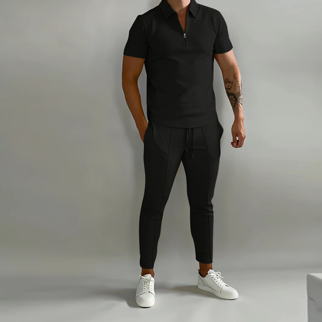 De afbeelding toont een man die een zwart, stijlvol trainingspak draagt van het merk Opestico. Het pak heeft een moderne snit, met een korte rits in de hals en een strakke pasvorm. De man draagt het pak in combinatie met witte sneakers, wat zorgt voor een casual, maar toch chique uitstraling. Dit trainingspak biedt zowel comfort als stijl, perfect voor sportieve en informele gelegenheden. De minimalistische uitstraling maakt het een veelzijdige keuze voor elke gelegenheid.