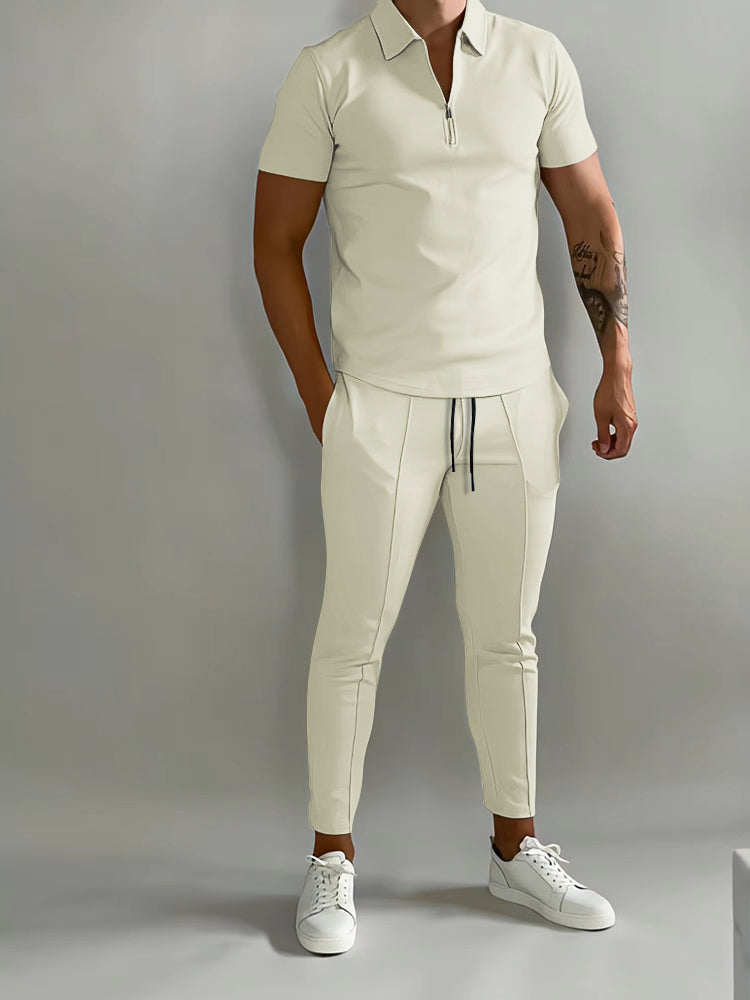 De afbeelding toont een man die een stijlvol trainingspak draagt van het merk Opestico. Het pak is wit met een korte rits in de hals en heeft een moderne, strakke pasvorm. De man combineert het trainingspak met witte sneakers voor een casual, maar toch chique look. Het kledingstuk benadrukt zowel comfort als stijl, perfect voor zowel sportieve als informele gelegenheden. Het strakke ontwerp en de minimalistische uitstraling maken het een veelzijdige keuze voor elke garderobe.
