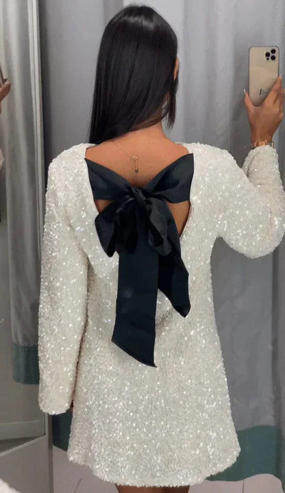 odel draagt de Ophira Dreamwear Elsa Glitter Jurk, een sprankelende witte jurk met een elegante zwarte strik aan de achterkant, terwijl ze zichzelf in de spiegel bewondert.