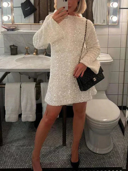 Model in de Ophira Dreamwear Elsa Glitter Jurk in wit, bedekt met glinsterende pailletten, terwijl ze poseert in een badkamer met een Chanel tas en zwarte hakken, met een spiegelreflexie in de achtergrond.