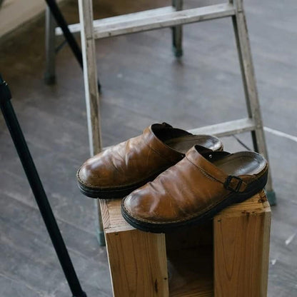 Bruine leren Paxano slippers geplaatst op een houten blok in een rustieke omgeving. De comfortabele en stijlvolle slippers hebben een verstelbare band, perfect voor dagelijks gebruik.