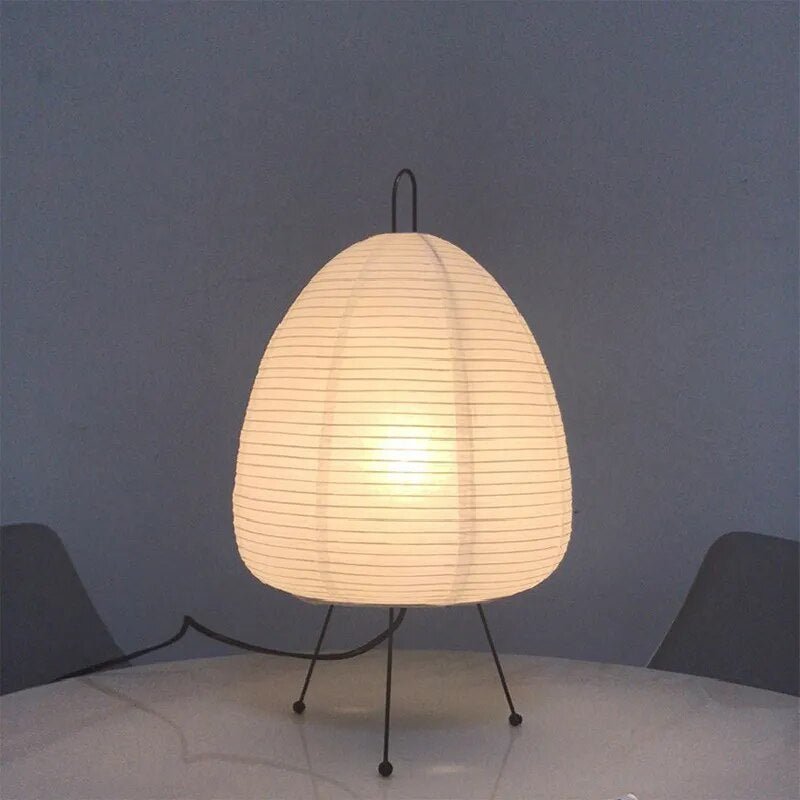 De Prana lamp, met een elegante papieren lampenkap in een organische, bolvormige vorm, rust op dunne, zwarte metalen poten. Het zachte licht creëert een gezellige en verfijnde sfeer, waardoor het de perfecte keuze is voor elke moderne interieur.