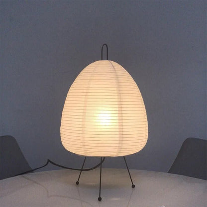 De Prana lamp, met een elegante papieren lampenkap in een organische, bolvormige vorm, rust op dunne, zwarte metalen poten. Het zachte licht creëert een gezellige en verfijnde sfeer, waardoor het de perfecte keuze is voor elke moderne interieur.