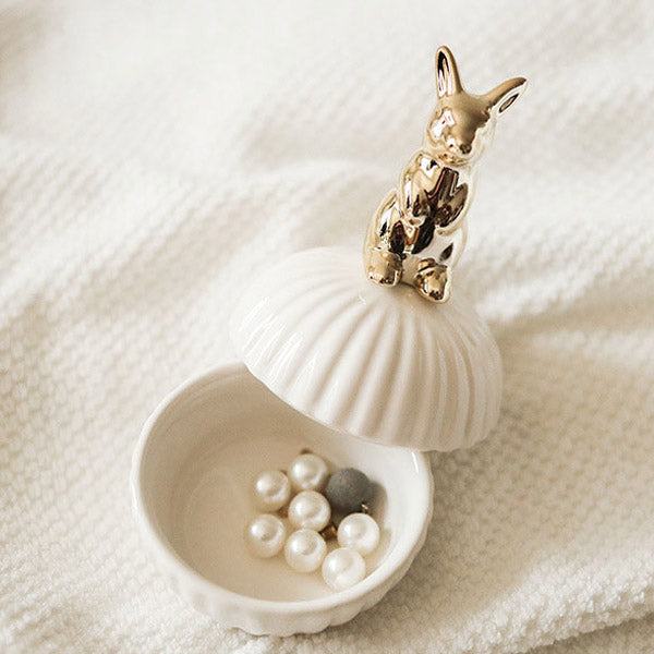 Een elegante opbergpot in de vorm van een schelp met een gouden konijn als deksel. De pot is geopend en bevat een paar parels. Het verfijnde ontwerp maakt het een stijlvolle en charmante opslagoplossing voor kleine voorwerpen.