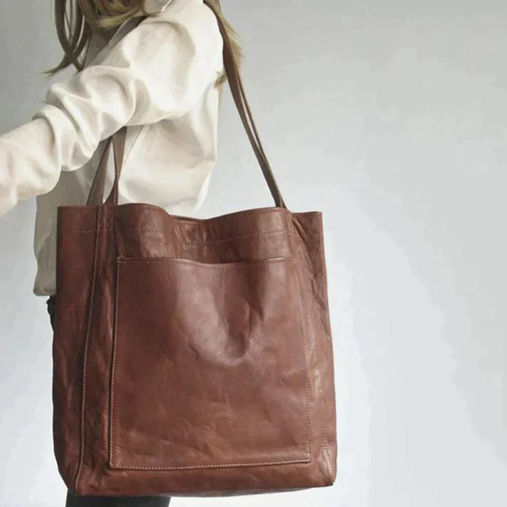 Een vrouw draagt een grote bruine leren schoudertas met een eenvoudige, minimalistische uitstraling. De tas heeft een ruime hoofdcompartiment met een zichtbare voorzak, en de lange hengsels maken het gemakkelijk om de tas over de schouder te dragen.