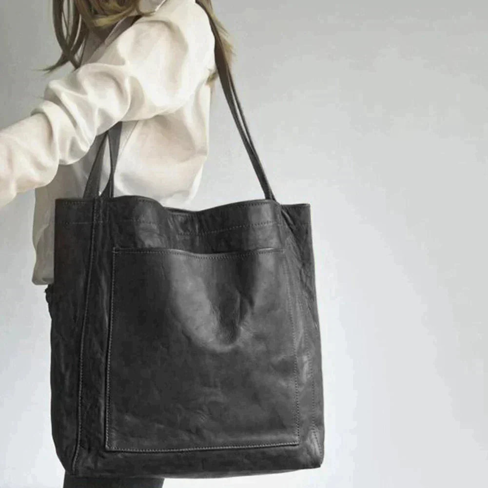 Een vrouw draagt een grote zwarte leren schoudertas met een minimalistisch ontwerp. De tas heeft een ruime hoofdcompartiment en een voorzak, ideaal voor dagelijks gebruik. De lange hengsels maken het gemakkelijk om de tas over de schouder te dragen.